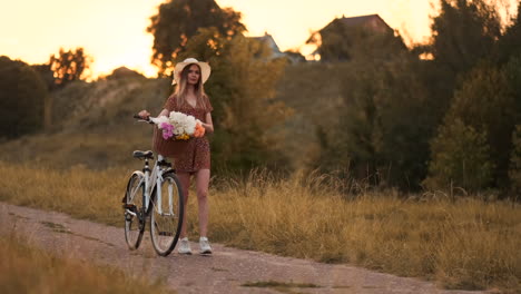 Young-beautiful-blonde-girl-in-summer-in-dress-and-hat-walking-on-road-with-bike-and-flowers-in-slow-motion.